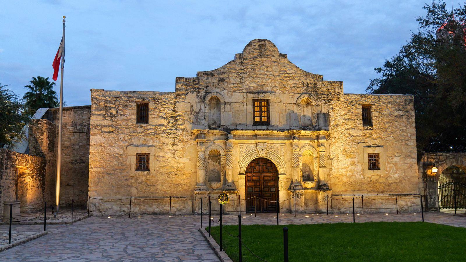 The Alamo lit at dusk with a Texas flag flying beside it