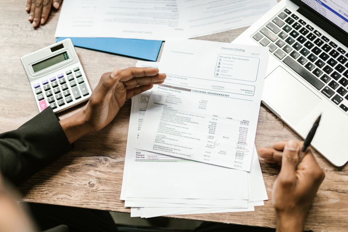 A bank statement held above a wooden desk beside a calculator and a laptop