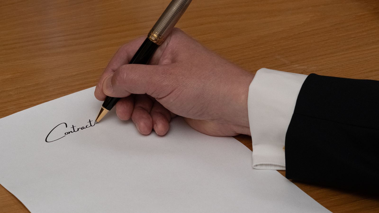 A hand signing a printed contract at a wooden desk